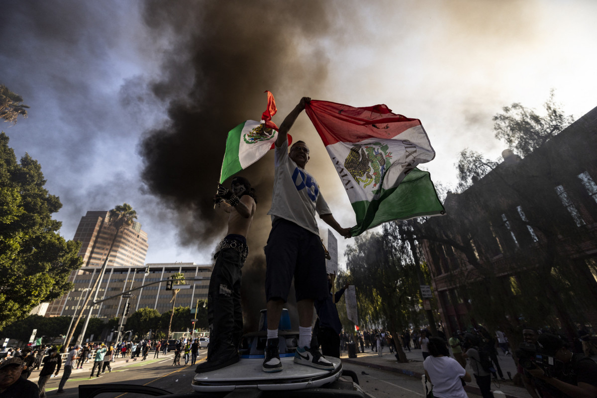 Manifestantes agitam bandeiras mexicanas do alto de um carro destruído, ao lado de veículos Waymo em chamas, enquanto se chocam com as forças da lei nas ruas ao redor do prédio federal durante um protesto após operações federais de imigração em Los Angeles, Califórnia, em 8 de junho de 2025. O presidente dos EUA, Donald Trump, enviou 2.000 soldados em 7 de junho para lidar com os crescentes protestos contra as operações de imigração na região de Los Angeles, uma ação que o governador do estado classificou como 